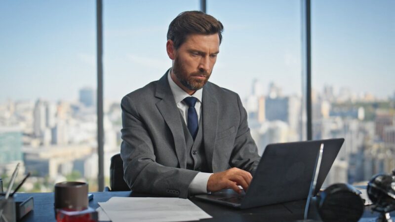 A focused attorney in a suit working on a laptop at a desk with a cityscape visible through large windows