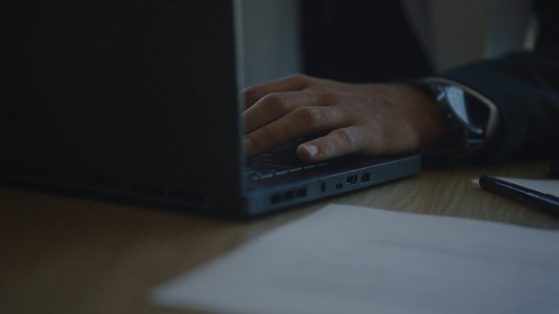 Close-up of a professional’s hand working on a laptop at a desk, with documents and a pen nearby