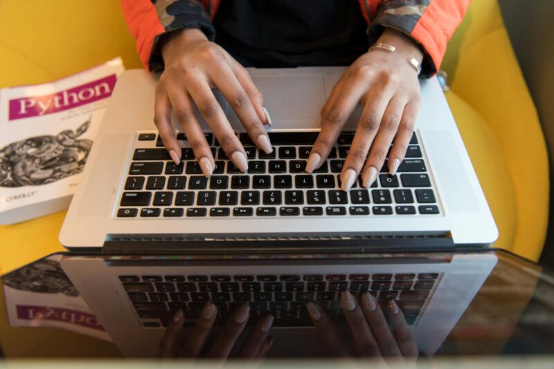 Hands typing on a laptop keyboard with a Python programming book in the background